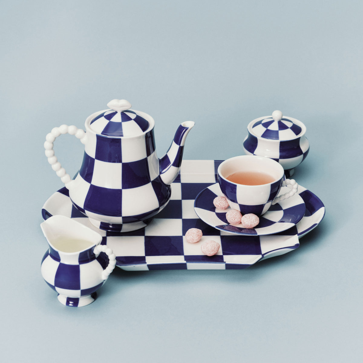 Het Serax Wouters & Hendrix Fête Des Bêtes Melkkannetje Blauw Wit wordt getoond als onderdeel van een blauw-wit geblokt Serax theeservies, gerangschikt met roze snoepjes op het schoteltje tegen een lichtblauwe achtergrond.