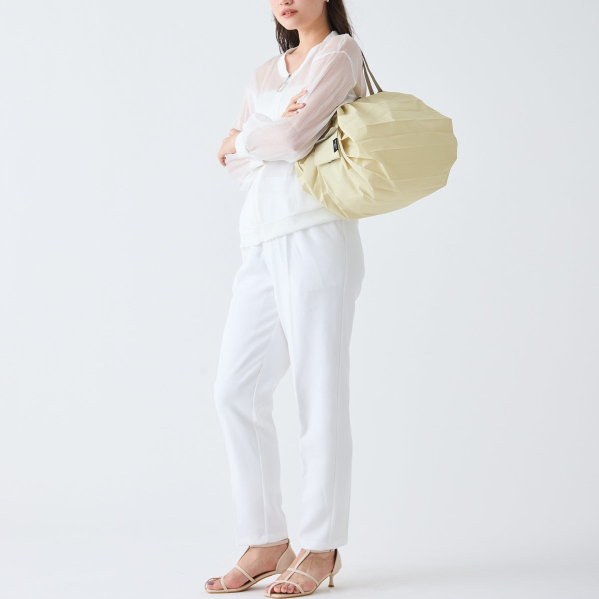 Een vrouw staat tegen een effen witte achtergrond en draagt een transparante witte blouse en broek. Ze draagt de Shupatto Opvouwbare Boodschappentas Cashewnoten Beige M over haar schouder met haar armen gekruist.