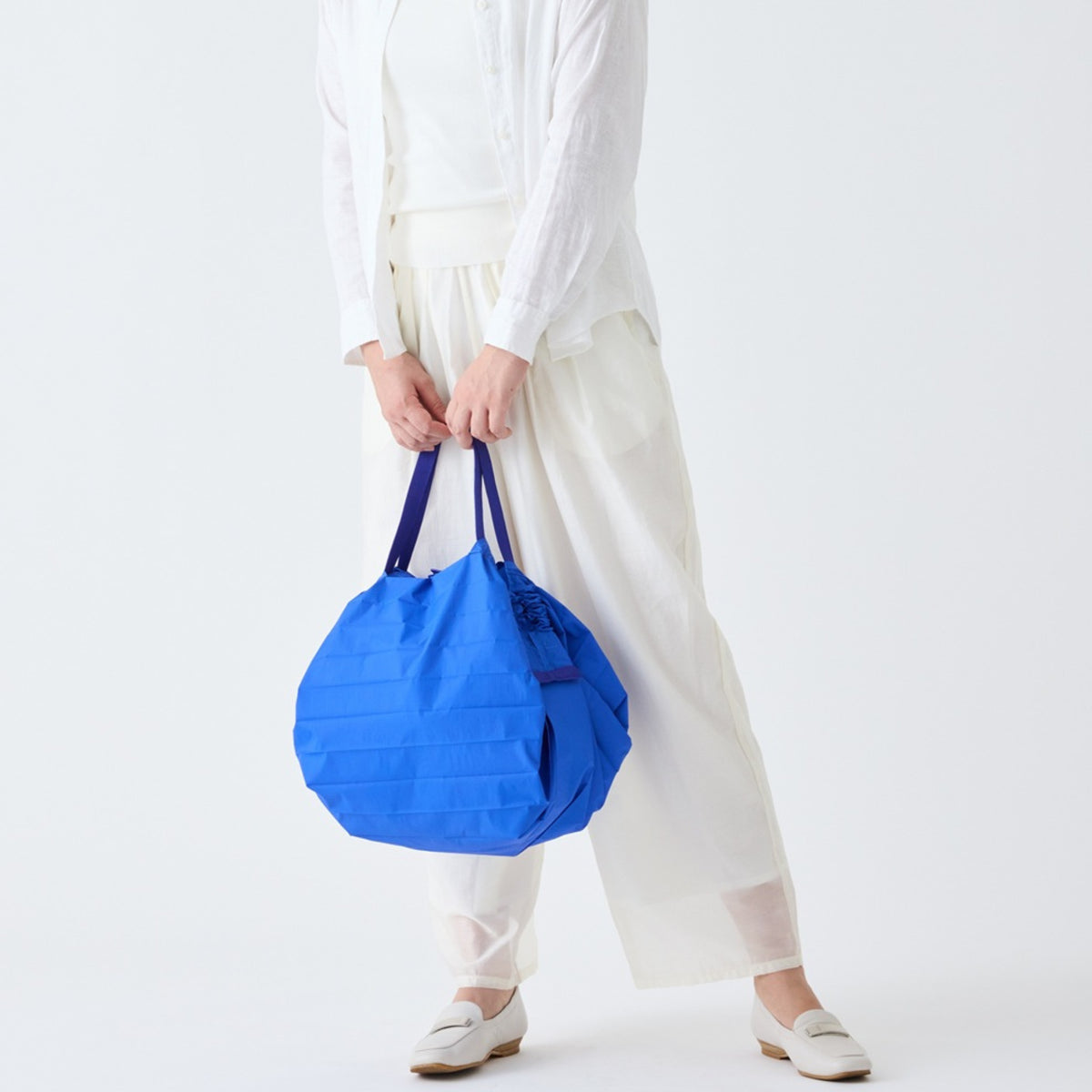 Een persoon in losse witte kleding en schoenen houdt een grote Shupatto Opvouwbare Boodschappentas Clear Sky Blauw M met donkere hengsels van Shupatto vast. De effen witte achtergrond en het onzichtbare gezicht van de persoon benadrukken de tas.