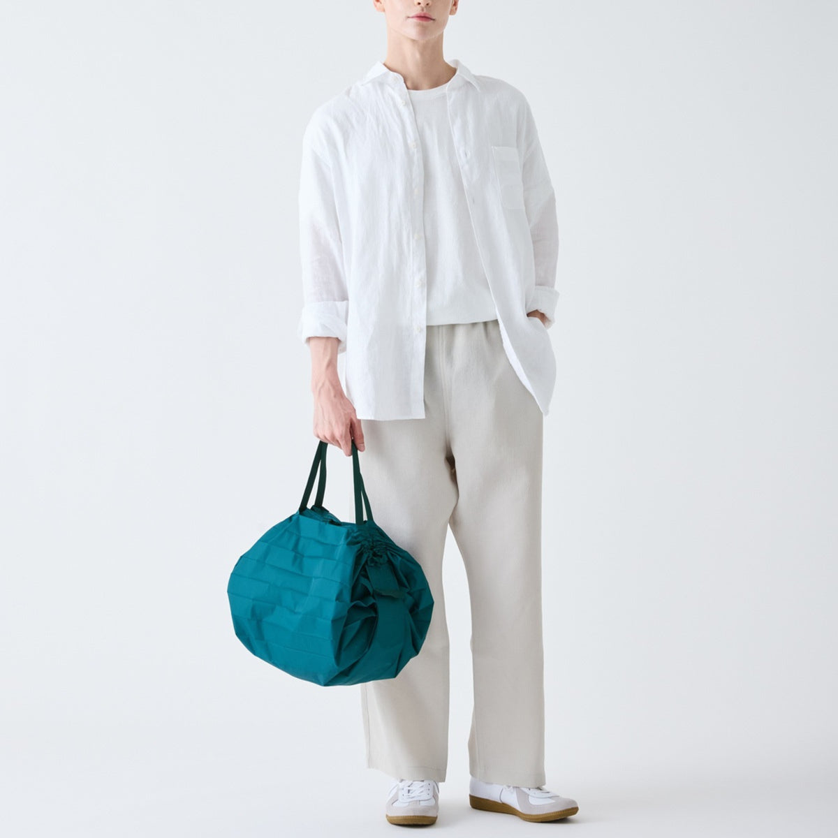 Een persoon in een wit t-shirt en lichtbeige broek houdt de Shupatto Opvouwbare Boodschappentas Donkergroen M vast en maakt zijn minimalistische look af met witte sneakers. Deze praktische, lichtgewicht tas van Shupatto is perfect voor dagelijks gebruik.