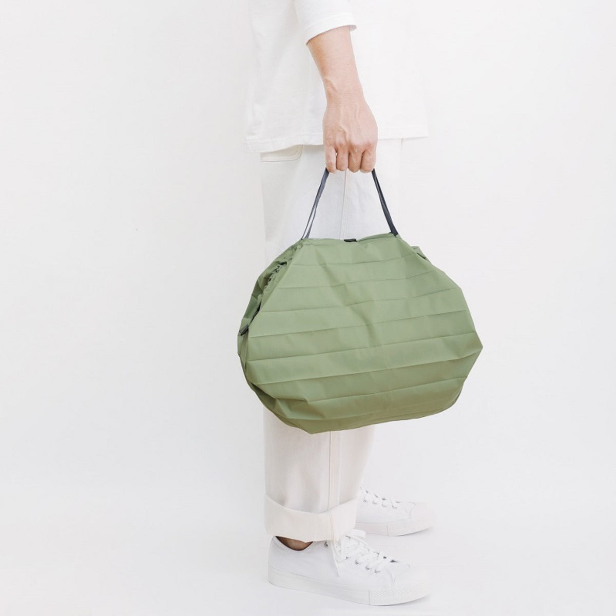 Een persoon in een witte broek, een wit shirt met lange mouwen en witte gymschoenen houdt de Shupatto Opvouwbare Boodschappentas Forest Groen M vast bij de zwarte handvatten tegen een effen witte achtergrond.