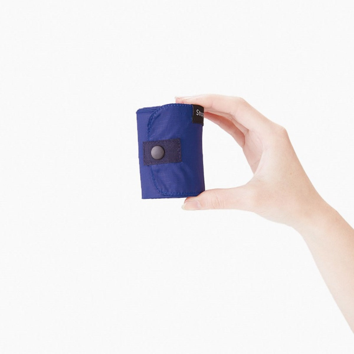 Een hand houdt een Shupatto Opvouwbare Boodschappentas Forest Groen M van Shupatto vast, compact opgerold in zijn etui met drukknoopsluiting, tegen een effen witte achtergrond.