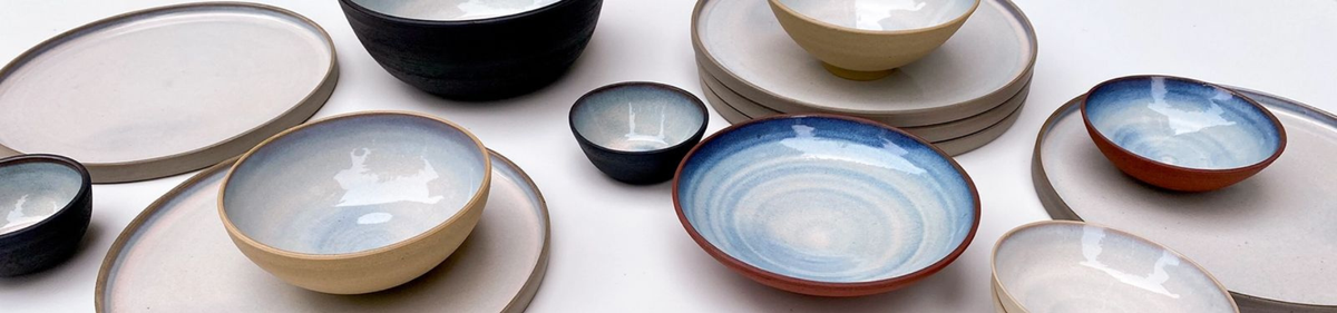 Verschillende handgemaakte keramische kommen en borden met neutrale en blauwe tinten zijn gerangschikt op een wit oppervlak, waarbij verschillende maten en vormen met een minimalistische, moderne esthetiek worden getoond.