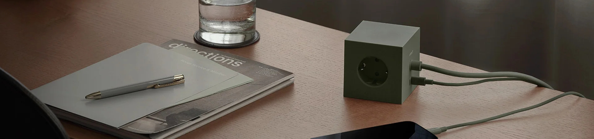 Een groene kubusvormige stekkerdoos op een houten bureau met twee snoeren aangesloten, naast een pen, papieren, een tijdschrift, een glas water en een smartphone.