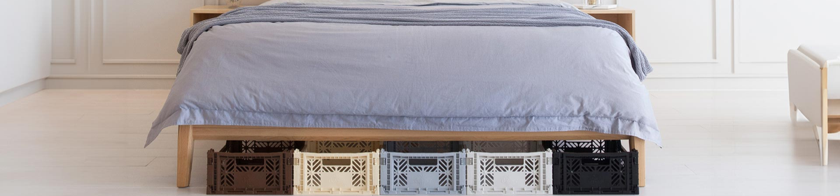 Een keurig opgemaakt bed met een blauwgrijs dekbed staat boven vier plastic opbergkratten in zwart, bruin, wit en grijs, georganiseerd onder het houten bedframe in een lichte, minimale slaapkamer.
