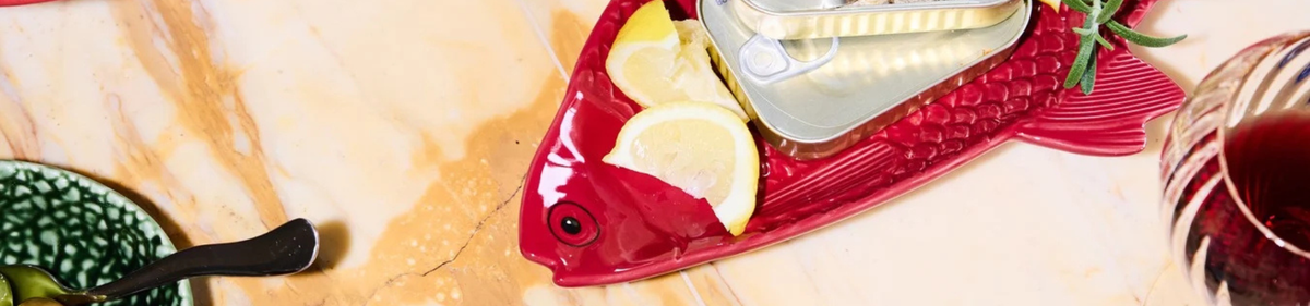 Op een rood visvormig bord liggen citroenschijfjes en een open blikje sardines. Het bord staat op een marmeren oppervlak naast een glas rode drank en een deel van een groene schaal met een lepel.