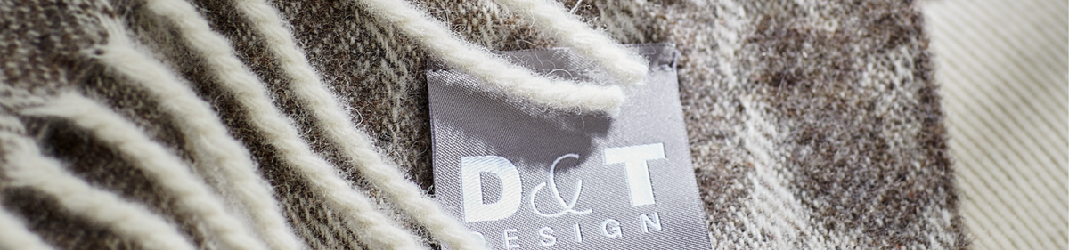 Close-up van een grijs stoffen label met D&T DESIGN tekst, bevestigd aan een wollen kledingstuk met zachte, crèmekleurige franjes en een bruin en beige structuurpatroon.