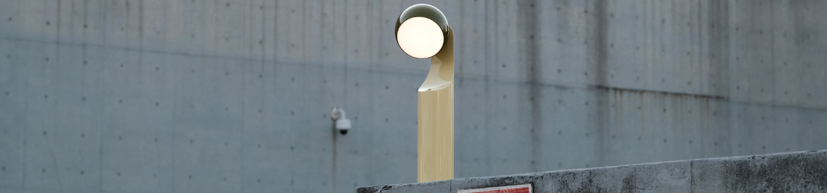 Een moderne, gele straatlantaarn met een ronde lampkop staat naast een betonnen muur; op de muur op de achtergrond is een beveiligingscamera gemonteerd.