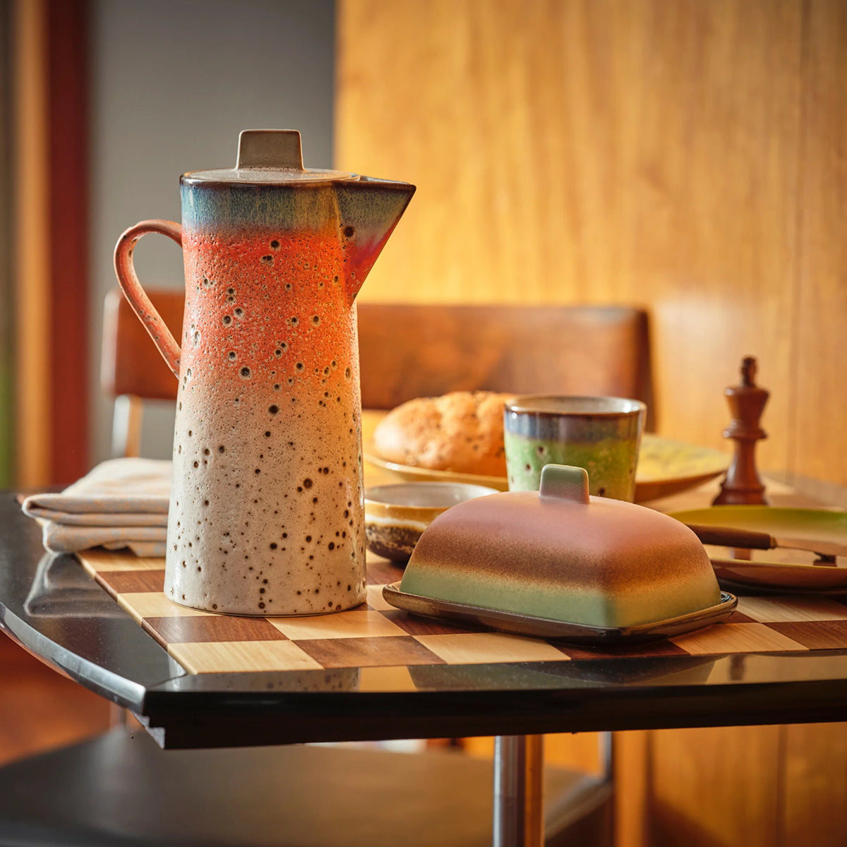 Een keramische kruik en botervloot met een gespikkeld, gradiëntglazuur staan op een ontbijttafel gedekt met brood, een kopje, servetten en een pepermolen in een gezellige, warm verlichte kamer.