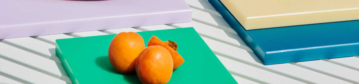 Twee oranje kaki's, waarvan één gehalveerd, rusten op een groen paneel. De scène bevat ook paarse, blauwe en beige panelen op een wit lattenoppervlak, waardoor een moderne, kleurrijke compositie ontstaat.