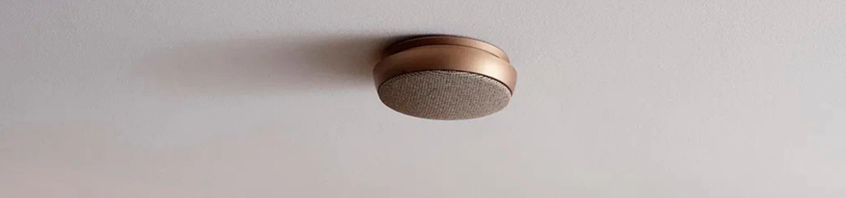 Een moderne, ronde rookmelder met een bronzen afwerking en een met stof bekleed hart is gemonteerd op een wit plafond.
