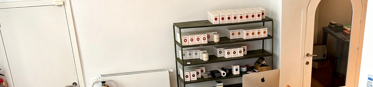 Een metalen plank met netjes gerangschikte koffieproductdozen en mokken staat tegen een witte muur bij een deuropening; een computer en kantoorbenodigdheden staan op een bureau op de voorgrond.