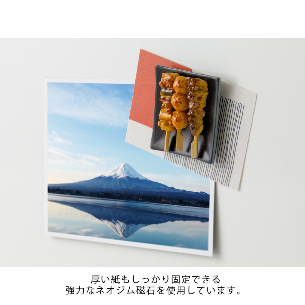 Een magneet van een yakitori-spies houdt een ansichtkaart van de berg Fuji en twee vellen papier tegen een wit oppervlak. Onder de afbeelding staat Japanse tekst.