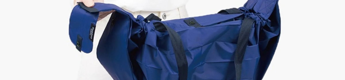 Een persoon in een witte outfit houdt een grote blauwe draagtas met zwarte riemen en trekkoorden aan de zijkant open, waardoor de ruime binnenkant zichtbaar wordt.