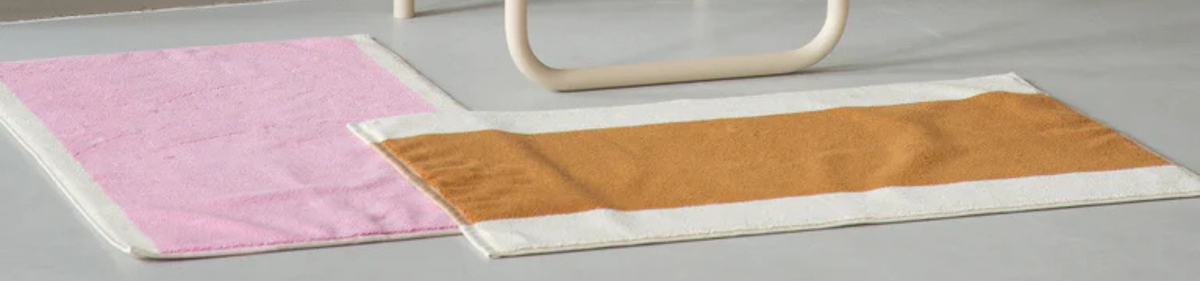 Op een grijze vloer liggen twee rechthoekige vloerkleden: het ene is effen roze en het andere heeft een brede taankleurige streep omzoomd door wit. Op de achtergrond is een gedeelte van gebogen, lichtgekleurd meubilair zichtbaar.