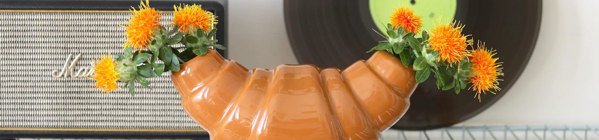 Een keramieken vaas in de vorm van een croissant houdt oranje bloemen vast aan beide uiteinden, met een vintage luidspreker en een vinylplaat op de achtergrond.