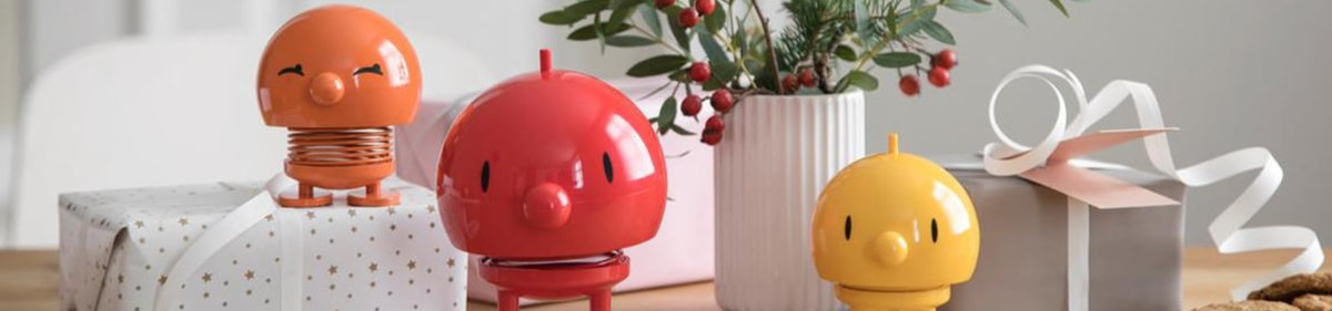 Drie kleurrijke, ronde lentefiguurtjes staan op een tafel met ingepakte cadeautjes en een vaas met groen en bessen. De beeldjes zijn oranje, rood en geel.
