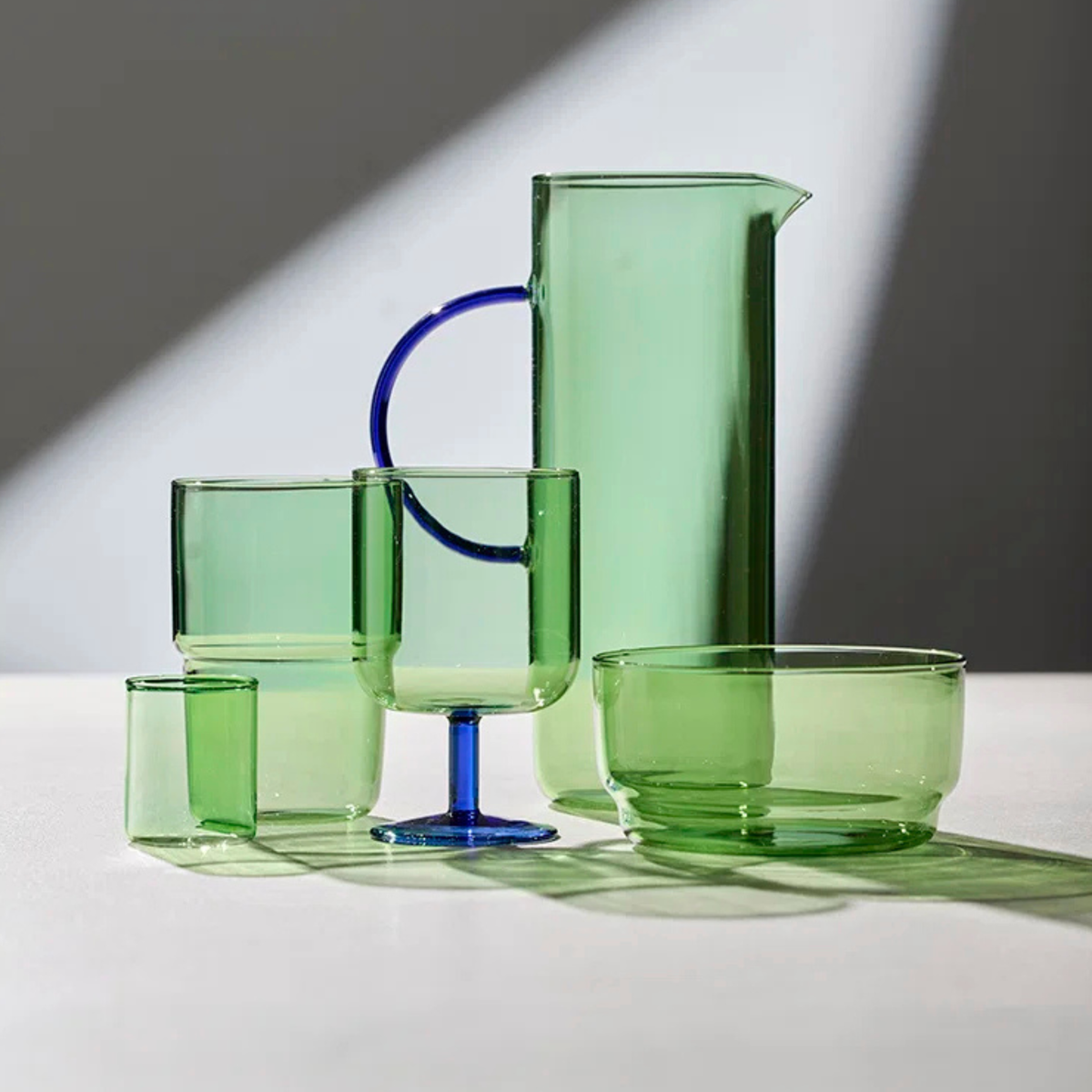 Een set elegant groen glaswerk, waaronder een kan, beker, twee bekers en een kom, staat op een wit oppervlak met dramatisch licht en schaduwen op de achtergrond.