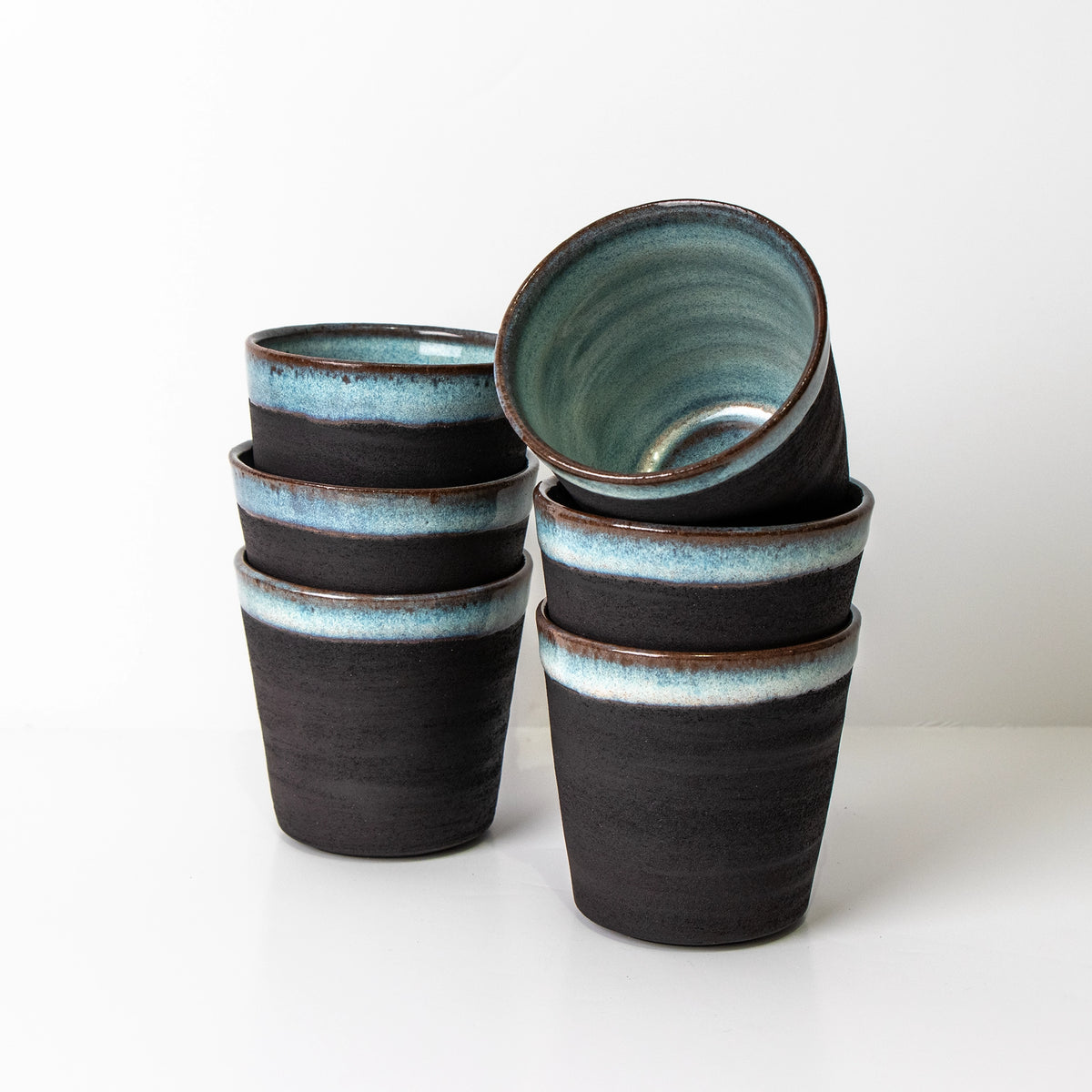 Zes AB Ceramics Beker Zwart Opaal 20 cl, met een matte zwarte buitenkant en blauw geglazuurde binnenkant, staan in twee stapels; één beker kantelt zichtbaar en laat het handgemaakt Antwerpen ontwerp zien.
