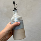Een hand houdt de AB Ceramics Olijfolie Fles Ribbel Wit Opaal 45 cl vast, met een geribbelde witte opalen bovenkant, gladde onderste helft en een metalen schenktuit, tegen een lichtgrijze wand met textuur.