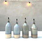 Vier AB Ceramics Olijfolie Fles Toffee Opaalgroen 55 cl dispensers staan op een rij op een wit oppervlak. De wazige achtergrond met lichtjes creëert een warme, gezellige gloed.