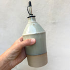 Een hand houdt de AB Ceramics Olijfolie Fles Toffee Opaalgroen 55 cl handgebakken keramiek met lichtgroen glazuur, naturel beige basis, metalen tuit en zwart rubberen stop tegen een effen lichte achtergrond.