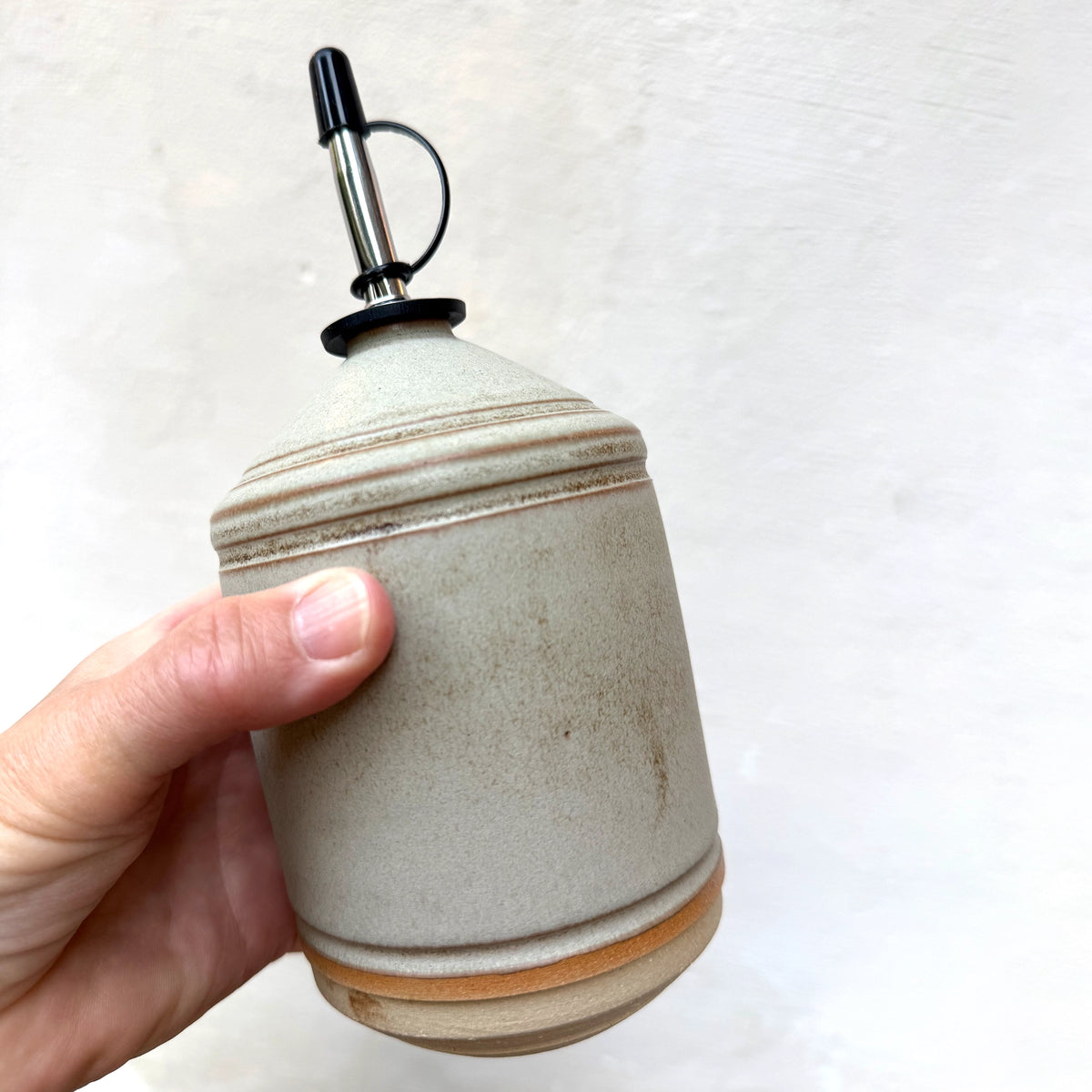 Een hand houdt de AB Ceramics Olijfolie Fles Wit Mos 55 cl met een metalen tuit en zwarte rubberen dop vast - een stijlvol AB Ceramics keukenaccessoire tegen een effen lichte achtergrond.