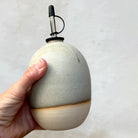 Een hand houdt de AB Ceramics Olijfolie Fles Wit Mos 60 cl vast, met een gladde, lichtgrijze ronde vorm met een metalen schenktuit en ongeglazuurde bodem die de natuurlijke klei onthult - een elegant keukenaccessoire.