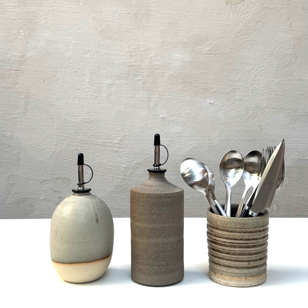 Drie AB Ceramics Olijfolie Fles Wit Mos 60 cl met zwarte tuiten staan op een wit oppervlak, naast een geribbelde beker met zilverwerk, alles tegen een lichtgrijze achtergrond-een tijdloze keuken accessoire set van AB Ceramics.