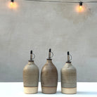 Drie AB Ceramics Olijfolie Fles 2 Wit Mos 55 cl staan op een rij op een witte ondergrond voor een grijze muur, met warme string lights erboven-een stijlvolle accessoire voor elke keuken.