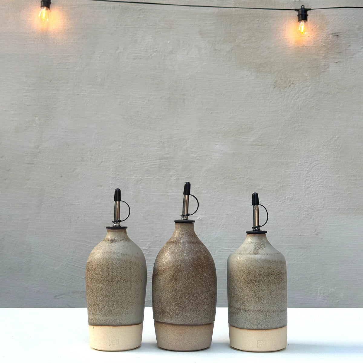 Drie AB Ceramics Olijfolie Fles 2 Wit Mos 55 cl staan op een rij op een witte ondergrond voor een grijze muur, met warme string lights erboven-een stijlvolle accessoire voor elke keuken.