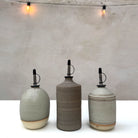 De AB Ceramics Olijfolie Fles Wit Mos 60 cl van AB Ceramics staat op een witte ondergrond. Boven de fles hangt een string met twee verlichte bollen tegen een lichtgrijze, getextureerde muur - een stijlvol keukenaccessoire.