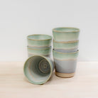 Zes AB Ceramics Bekers Wit Opaal/Lichtgroen van 20 cl zijn gestapeld op licht hout. Eén handgemaakte beker van AB Ceramics ligt horizontaal en toont het interieur met een fraaie swirl.