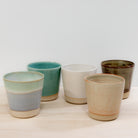 De AB Ceramics Beker Wit Opaal/Lichtgroen 20 cl, een uniek handgemaakte beker van wit steengoed, staat op een licht houten oppervlak tegen een effen witte achtergrond.