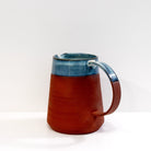 De AB Ceramics Karaf 5 Rood Opaal Blauw 100 cl heeft een handgegoten body van rode klei, een brede basis en opaalglazuur op de bovenste helft en het handvat, tegen een effen witte achtergrond.