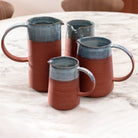 Vier AB Ceramics Karaf 6 Rood Opaal Blauw 50 cl kruiken met grote handgrepen en blauw opaalglazuur staan bij elkaar op een ronde tafel van wit marmer.