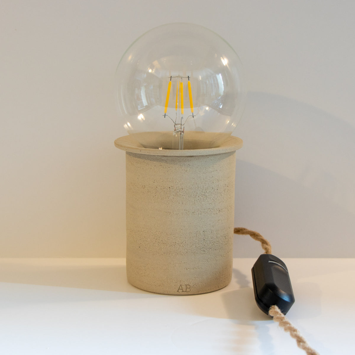 De AB Ceramics LED Lamp Dimmer is een minimalistische, handgemaakte lamp met een ronde heldere lamp en cilindrische beige keramische voet. De lamp heeft een bruin stoffen snoer en een zwarte inline schakelaar, gemaakt door AB Ceramics.