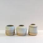 Drie kleine, ronde AB Ceramics Vaasje Wit Opaal vazen met glanzend lichtgrijs en beige opaalglazuur staan op een rij op een witte achtergrond. Het linker vaasje heeft horizontale ribbels; de andere zijn glad.