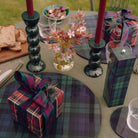 Op een buitentafel liggen geruite placemats, ingepakte cadeautjes, een 24 cm Addison Ross Bobbin Kandelaar in Racing Green met rode kaarsen, een vaas met wilde bloemen, glaswerk en een houten plank met sneetjes brood.