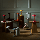 Een groep kleurrijke, moderne tafellampen, waaronder de Addison Ross Bobbin Lamp LED Oplaadbaar Kersenrood, hebben ronde lampvoeten en koepelvormige kappen. Ze staan op houten statieven tegen een wand met panelen in een zacht verlichte kamer.