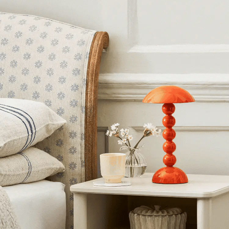 Op een nachtkastje naast het bed staan een Addison Ross Bobbin Lamp LED Oplaadbaar Oranje, een glas water, witte bloemen in een kleine vaas en een kaars. Gedessineerde kussens sieren het bed en het tafeltje staat tegen een witte muur.