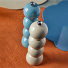 Twee Addison Ross Bobbin Zout-en Pepermolen Chambray 24 cm slijpmachines - een pastelblauwe, een gebroken witte - staan rechtop op een oranje gestructureerd oppervlak, met chambray en beige placemats op de achtergrond.