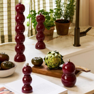 Een 35 cm kersenrood Addison Ross Bobbin zout-en pepermolen staat op een aanrecht met heirloomtomaten, artisjok, kruiden in potten, een open boek en een gestreept gordijn op de achtergrond.