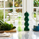 Twee stijlvolle Addison Ross Bobbin Zout-en Pepermolen Racing Groen 24 cm van Addison Ross staan op een licht aanrecht in de buurt van verse groenten, een glas water, een snijplank en zonovergoten ramen met uitzicht op de tuin.