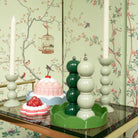 Een decoratieve tafel met Addison Ross Bobbin kandelaars met witte kaarsen, een groene Addison Ross Bobbin Zout-en Pepermolen Racing Groen 24 cm, twee desserts, alles tegen vogel- en takkenbehang met een vogelkooi.