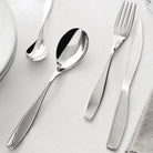 Een Alessi Duna Bestek 24-delig servies met Italiaans design staat op een wit tafelkleed naast een bord en servet.
