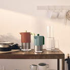 De Alessi Vite Koffiemaker Saliegroen 3-Kops van Alessi staat op een rustieke houten tafel met mokken, glazen en een karaf. Drie witte kopjes hangen aan een nabijgelegen muur. Perfect voor inductiegebruik en stijlvolle koffiemomenten.
