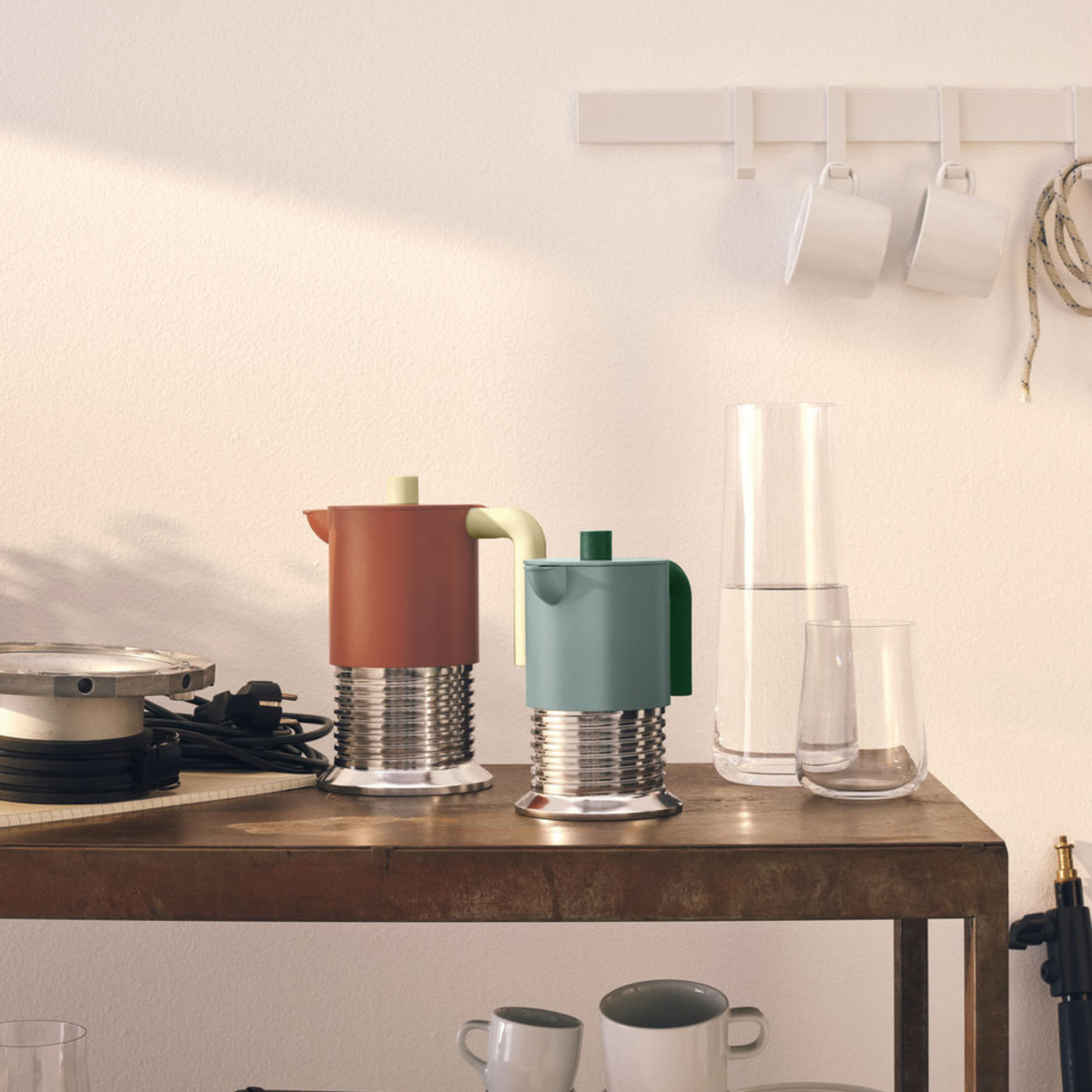 De Alessi Vite Koffiemaker Saliegroen 3-Kops van Alessi staat op een rustieke houten tafel met mokken, glazen en een karaf. Drie witte kopjes hangen aan een nabijgelegen muur. Perfect voor inductiegebruik en stijlvolle koffiemomenten.