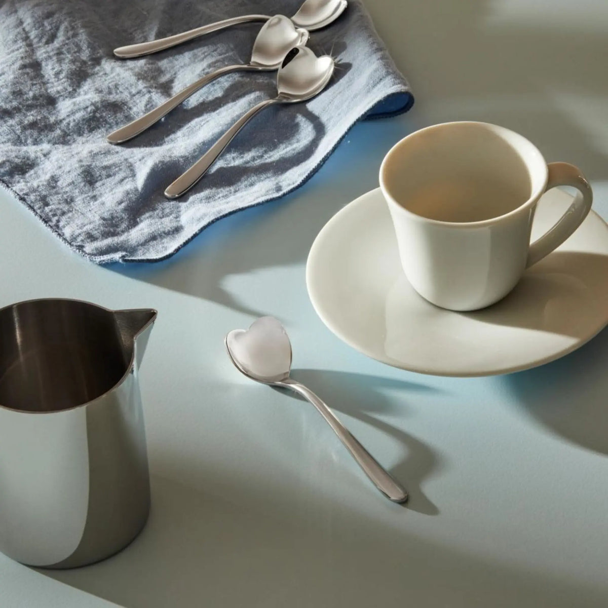 Een witte kop en schotel op een lichtblauw oppervlak staan naast een roestvrijstalen melkkan, een lepel en een blauwe doek met daarop vier Alessi Big Love Koffielepels 4 st. van Alessi. Helder licht werpt zachte schaduwen.