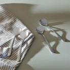 De Alessi Big Love Koffielepels 4 st. van Alessi staan op een licht oppervlak - twee op een blauw-wit gestreepte doek en twee ernaast - en werpen elk zachte schaduwen.